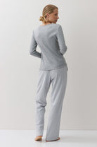 A woman stands barefoot against a plain white studio background, viewed from the back, wearing a light gray long-sleeve top and matching white-and-blue checkered pajama pants.