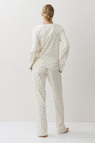 A woman stands with her back to the camera wearing a matching long-sleeve pajama top and pants set in a warm off-white fabric with a small, repeating floral print.