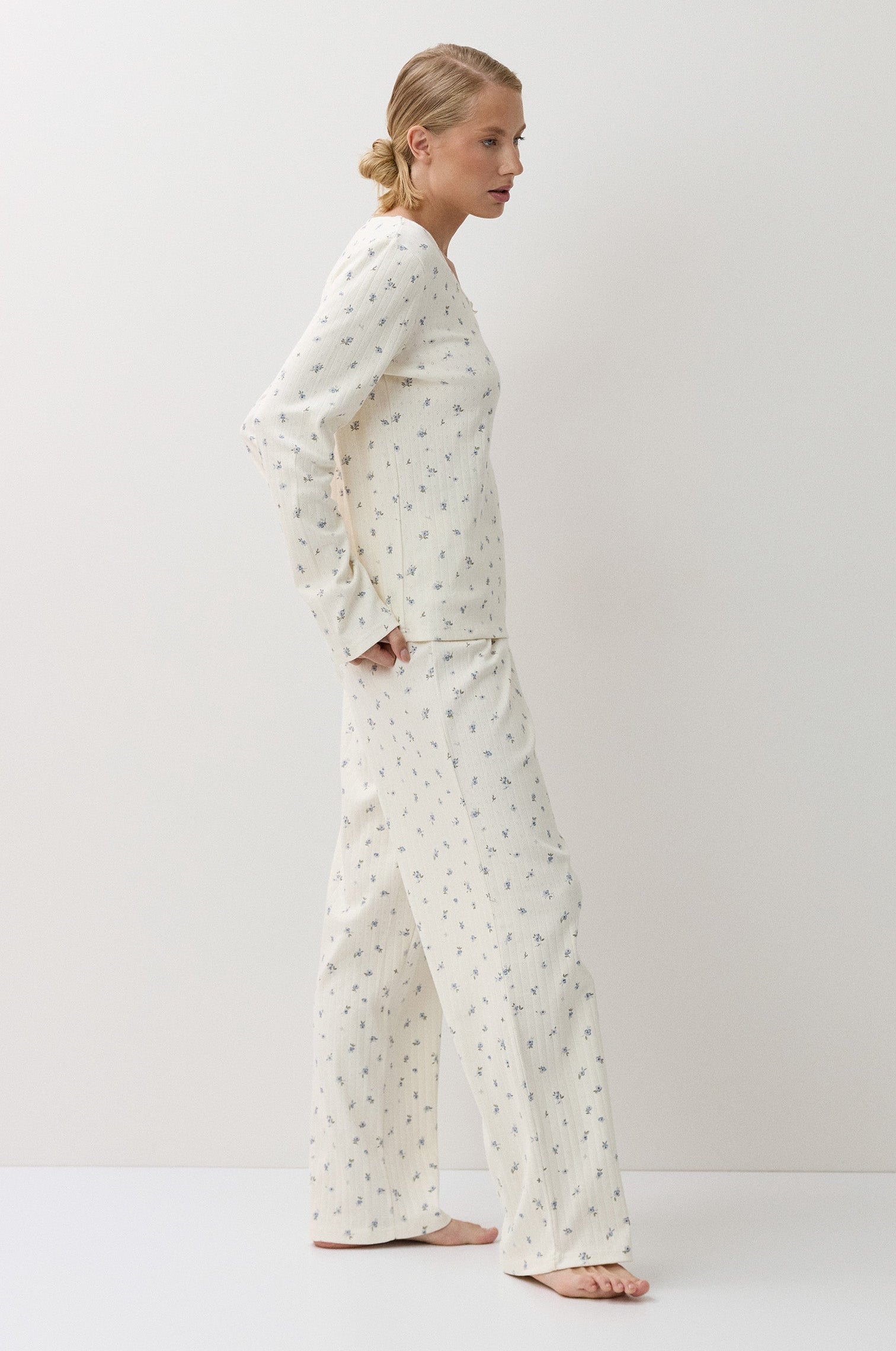 A woman stands in profile wearing a matching long-sleeve pajama top and wide-leg pants set in a warm off-white fabric with a small, repeating green floral print. The photo is taken in a minimalist studio setting with a light white background.