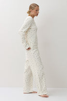 A woman stands in profile wearing a matching long-sleeve pajama top and wide-leg pants set in a warm off-white fabric with a small, repeating green floral print. The photo is taken in a minimalist studio setting with a light white background.