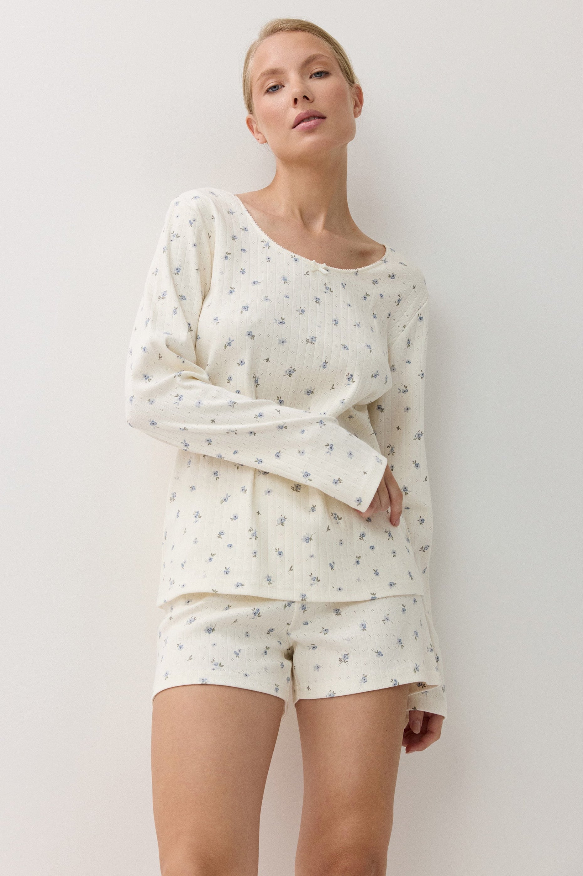 A woman modeling a two-piece pajama set consisting of a long-sleeve top and matching shorts, both in an off-white color with a small blue floral pattern, set against a plain white  background.