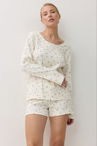 A woman modeling a two-piece pajama set consisting of a long-sleeve top and matching shorts, both in an off-white color with a small blue floral pattern, set against a plain white  background.