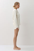 A woman stands in profile wearing a matching long-sleeve pajama top and shorts set in a warm off-white fabric with a small, scattered floral print. The image is shot in a minimalist studio setting with a warm, off-white background.