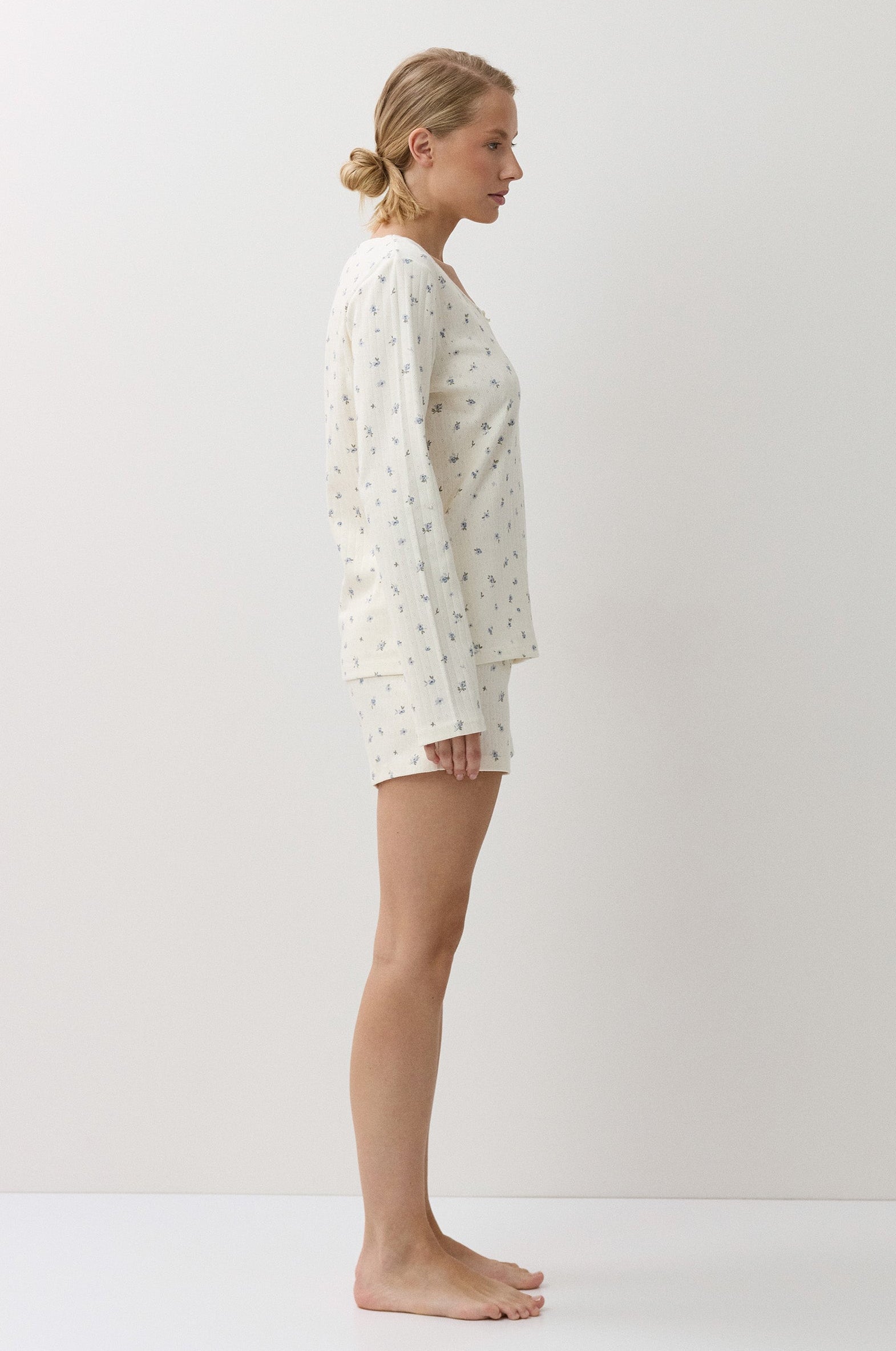 A woman stands in profile wearing a matching long-sleeve pajama top and shorts set in a warm off-white fabric with a small, scattered floral print. The image is shot in a minimalist studio setting with a warm, off-white background.