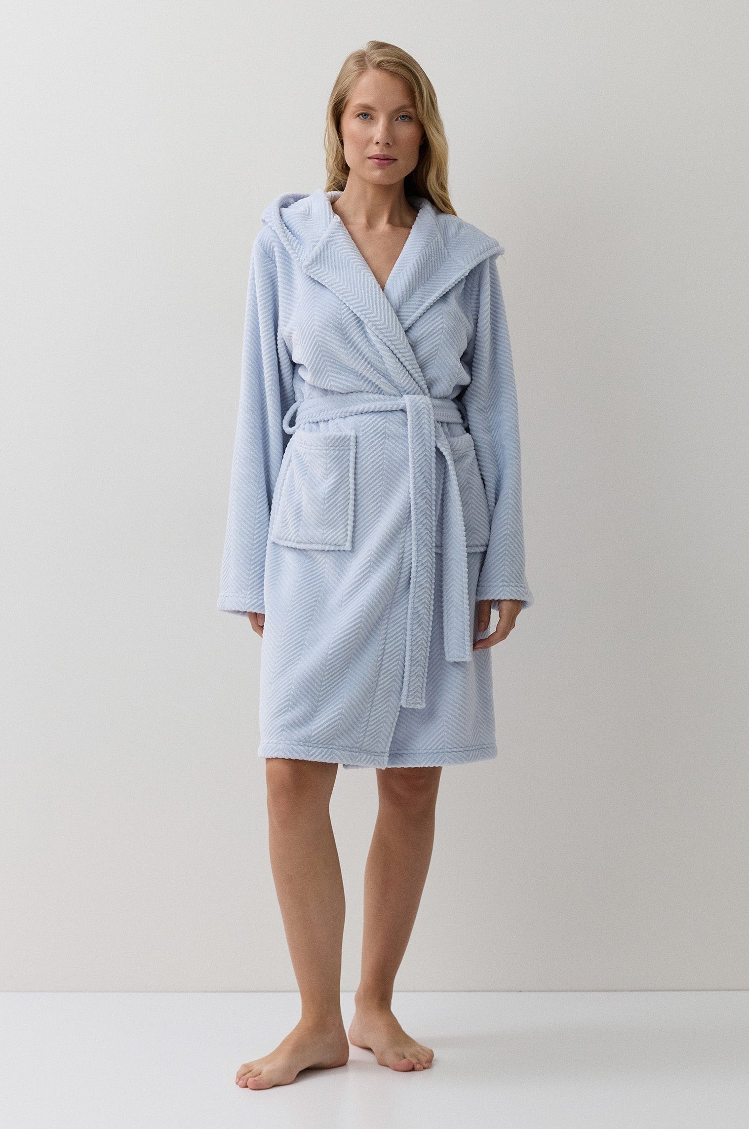 A woman modeling a light blue hooded bathrobe with a tie belt and front patch pocket, standing against a plain off-white studio background.