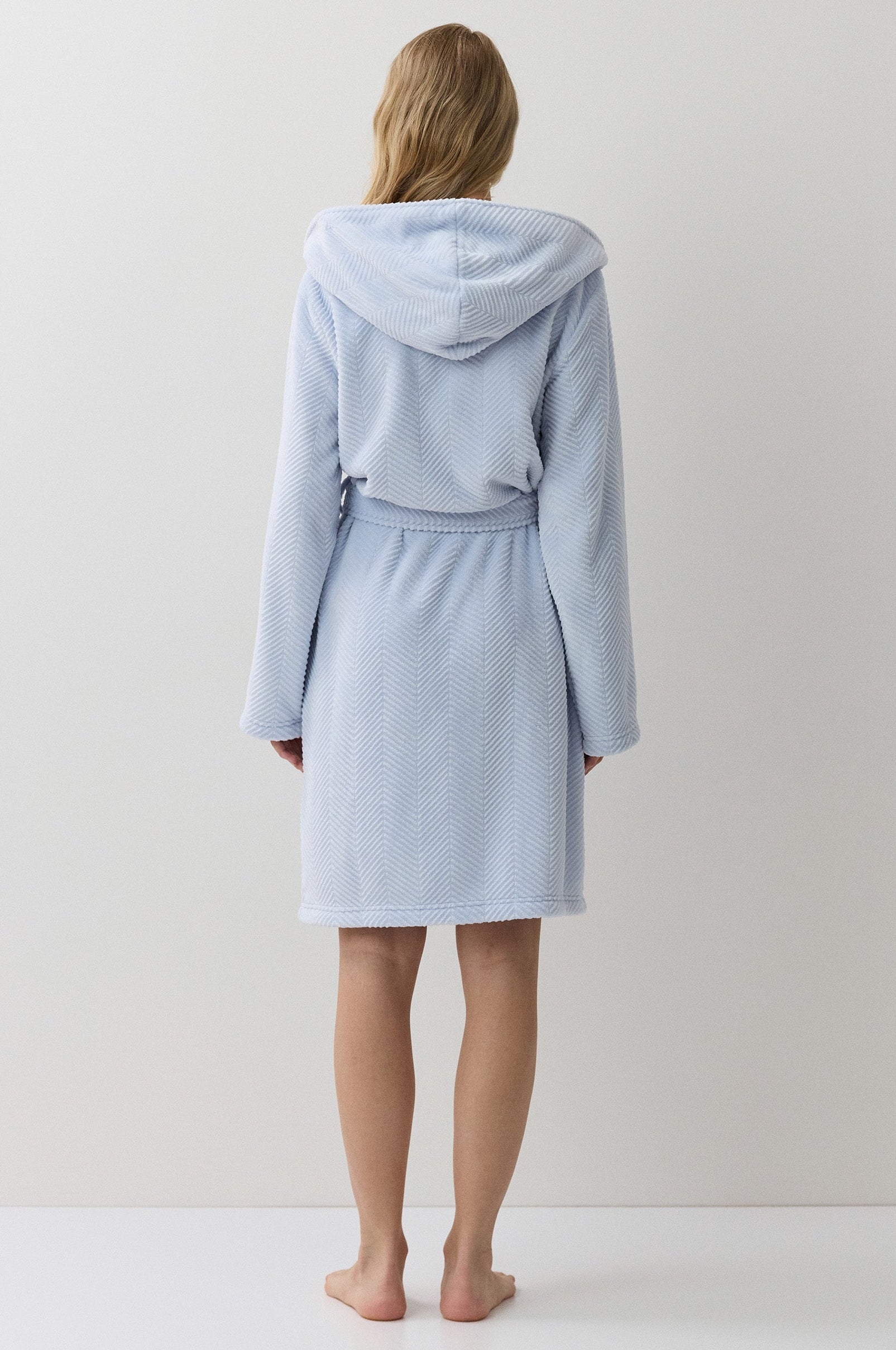 A woman stands with her back to the camera wearing a light blue hooded bathrobe against a plain white studio background.