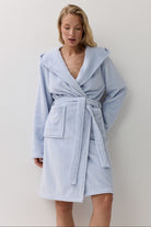 A woman wearing a light blue textured bathrobe with a hood, waist tie, and front patch pockets, standing against a plain white wall.