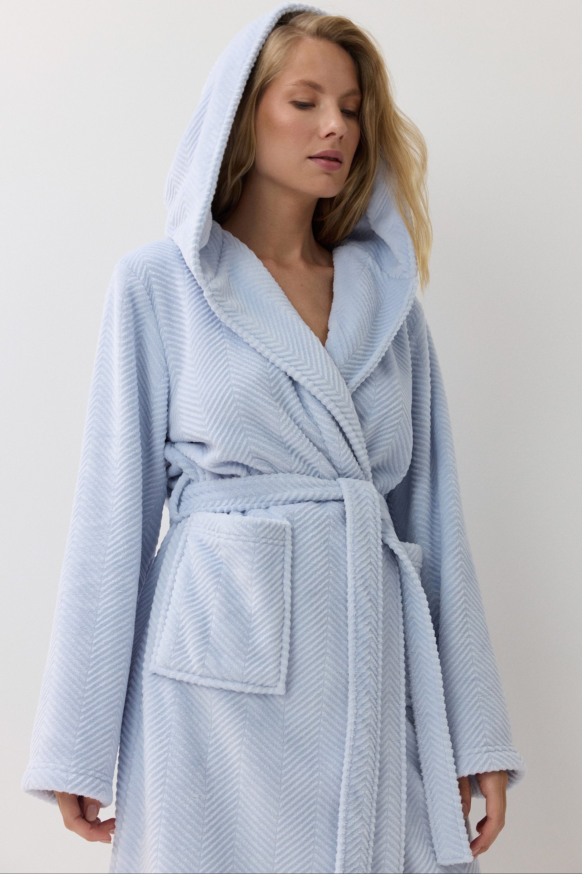 A woman wearing a light blue hooded bathrobe with a textured pattern, standing against a plain off-white background.