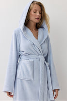 A woman wearing a light blue hooded bathrobe with a textured pattern, standing against a plain off-white background.