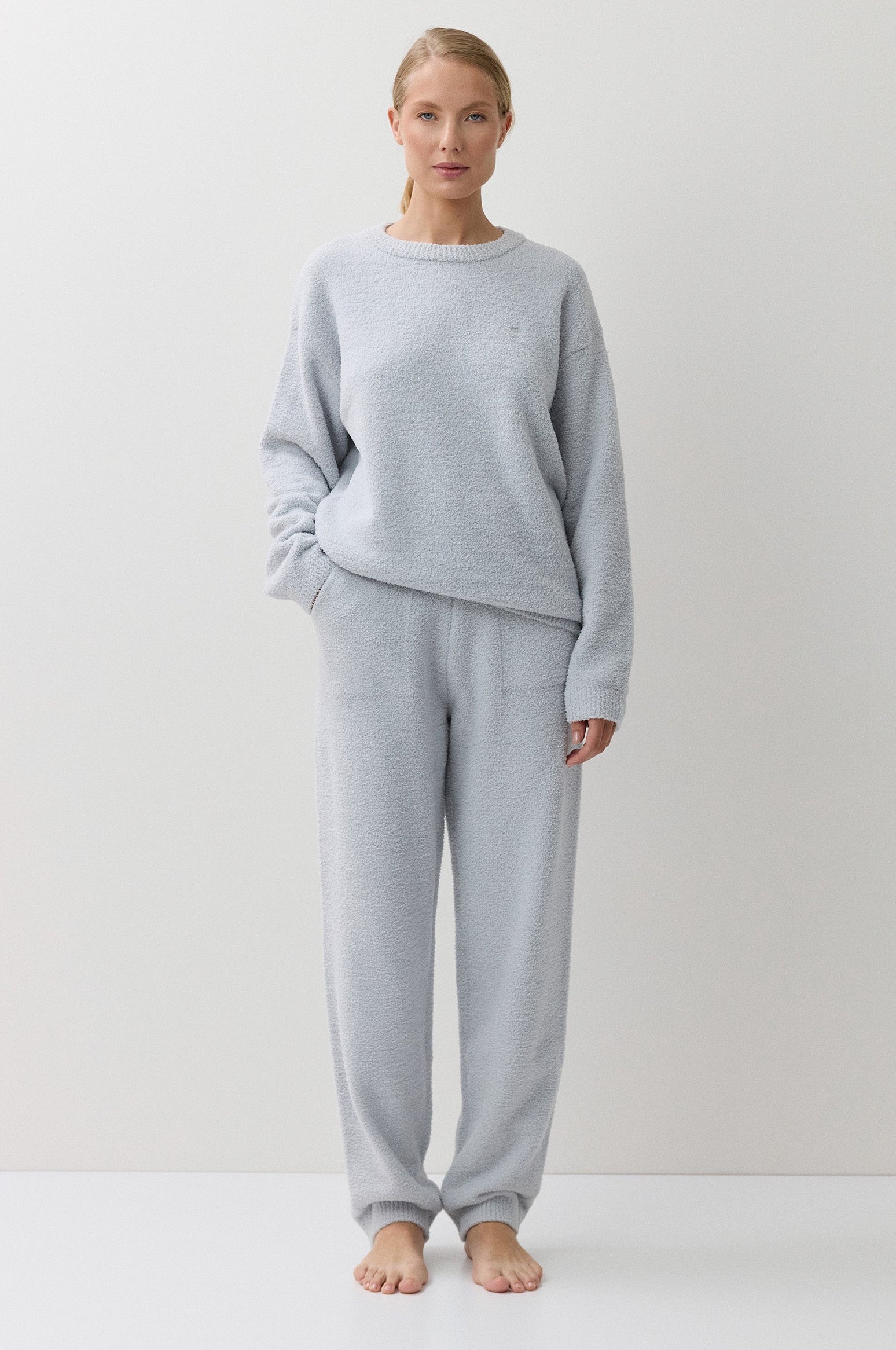 A woman stands barefoot against a plain white studio background, wearing a light blue long-sleeve sweatshirt and matching jogger-style pants.