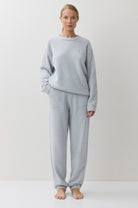 A woman stands barefoot against a plain white studio background, wearing a light blue long-sleeve sweatshirt and matching jogger-style pants.