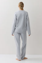 A woman stands with her back to the camera wearing a light blue lounge set consisting of a long-sleeve top and matching pants, set against a plain off-white studio background.