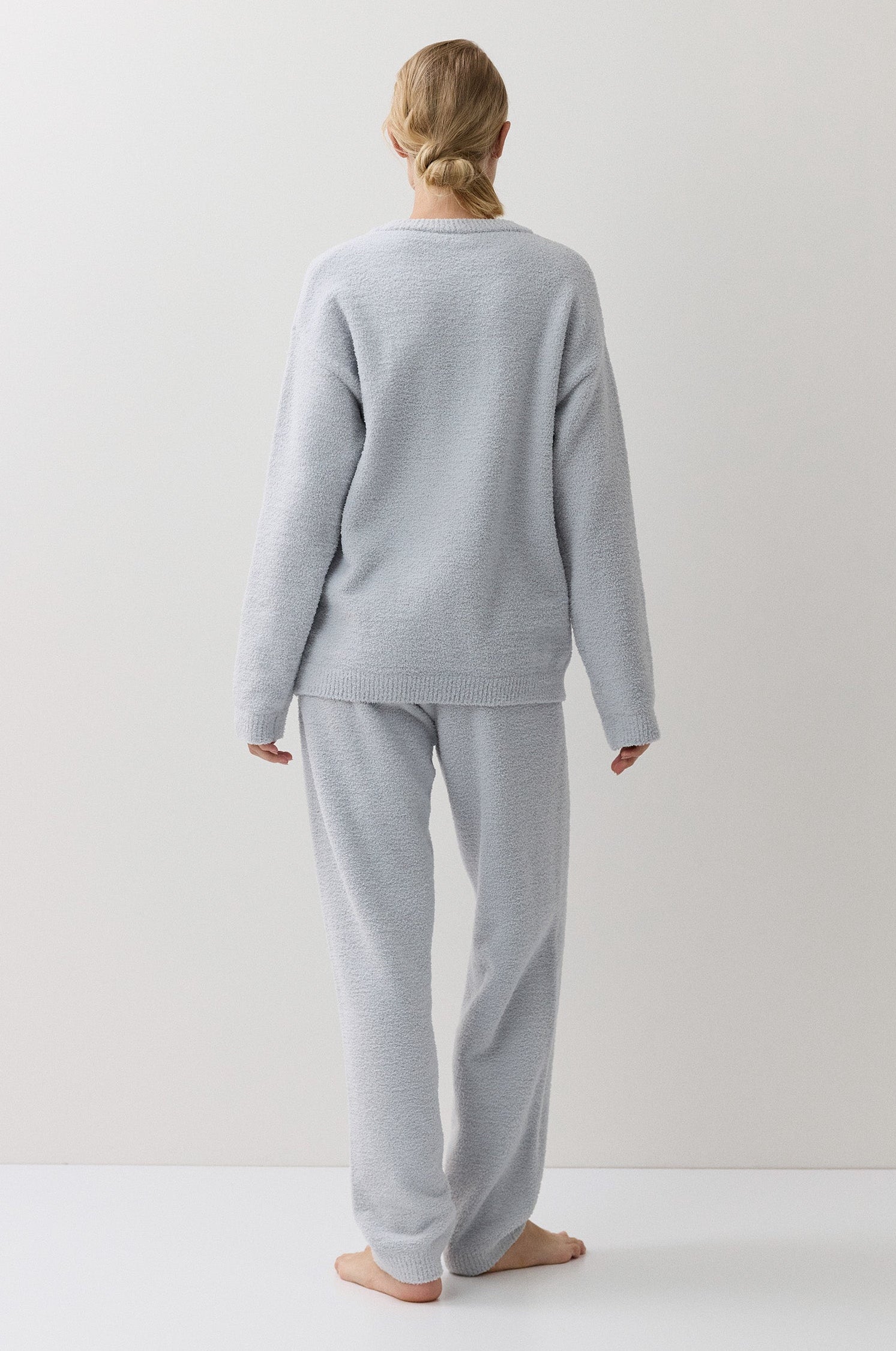 A woman stands with her back to the camera wearing a light blue lounge set consisting of a long-sleeve top and matching pants, set against a plain off-white studio background.
