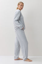 A woman stands barefoot in profile wearing a light blue two-piece lounge set consisting of a long-sleeve sweatshirt and matching wide-leg sweatpants against a plain white studio background.