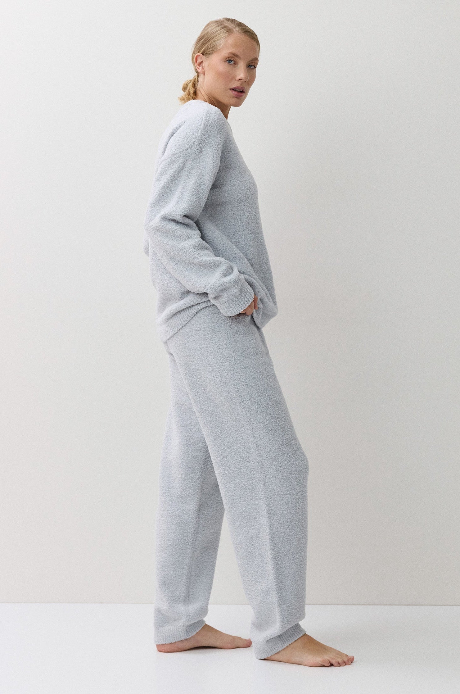 A woman stands barefoot in profile wearing a light blue two-piece lounge set consisting of a long-sleeve sweatshirt and matching wide-leg sweatpants against a plain white studio background.