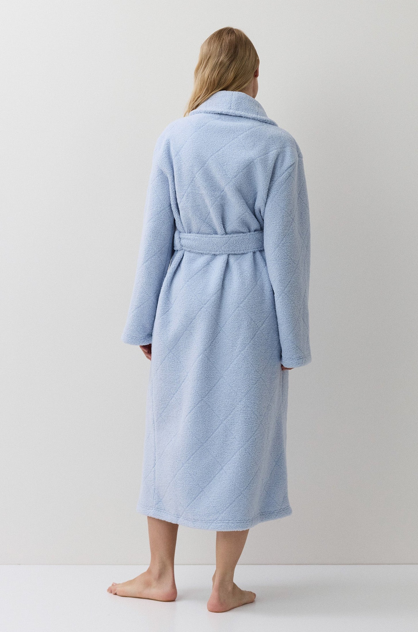 A woman stands with her back to the camera wearing a light blue bathrobe with a matching tie belt, set against a plain white studio background.