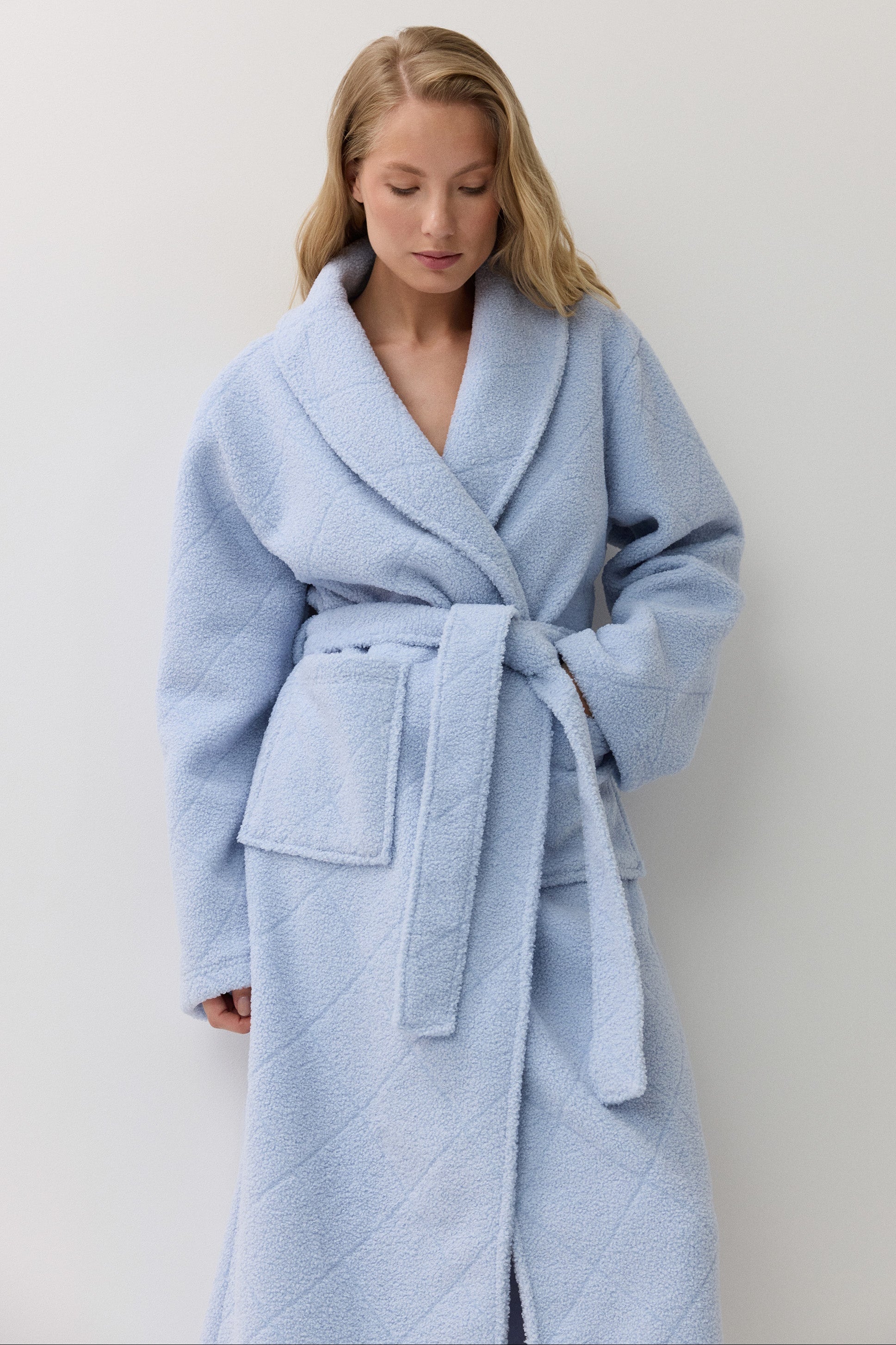 A woman wearing a light blue quilted bathrobe with a shawl collar and tie belt, standing against a plain light gray wall.