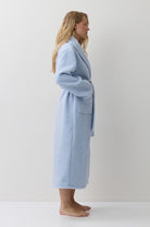 A woman stands in profile wearing a light blue bathrobe against a plain white studio background.