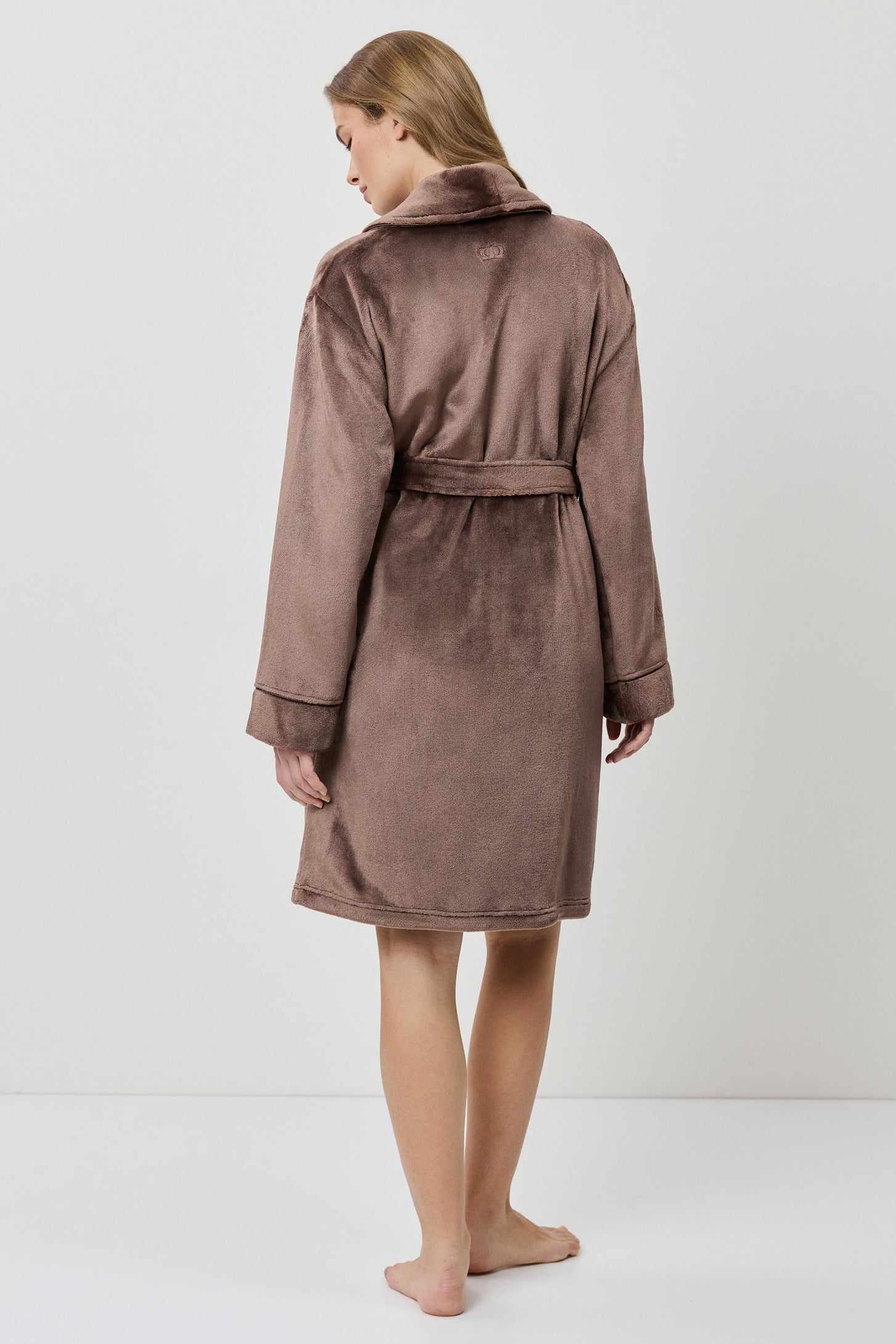 A woman wearing a plush, knee-length bathrobe from the back. The robe is a muted taupe-brown color with a soft, velvety texture and a matching waist tie.
