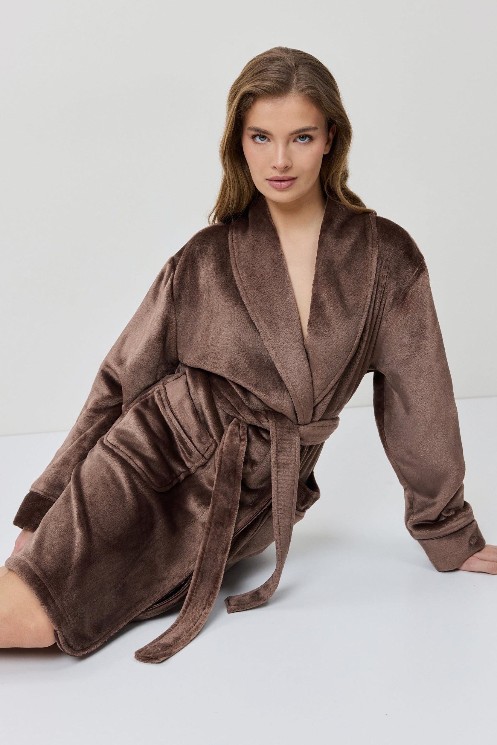 A woman wearing a plush, knee-length bathrobe from the back. The robe is a muted taupe-brown color with a soft, velvety texture and a matching waist tie.