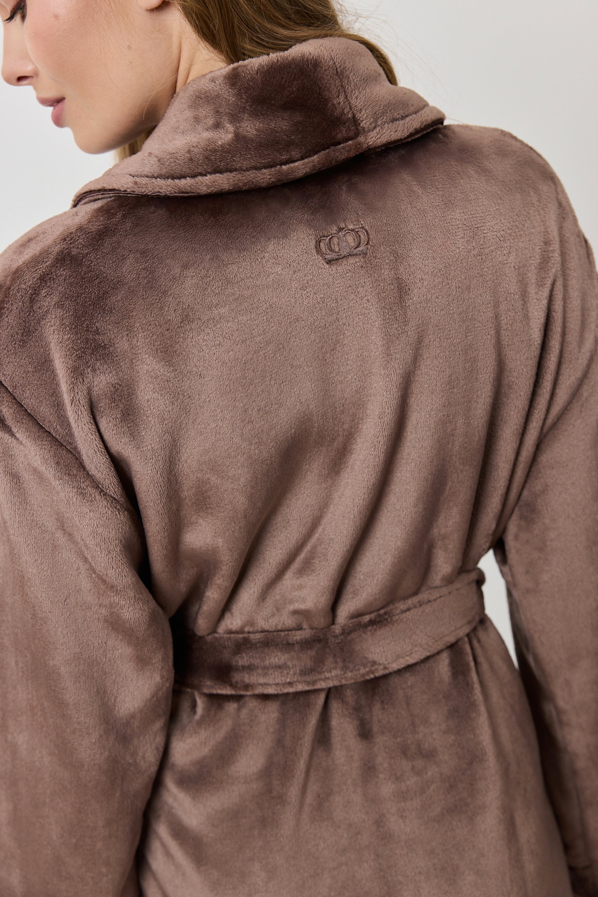 A woman wearing a plush, knee-length bathrobe from the back. The robe is a muted taupe-brown color with a soft, velvety texture and a matching waist tie.