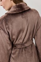A woman wearing a plush, knee-length bathrobe from the back. The robe is a muted taupe-brown color with a soft, velvety texture and a matching waist tie.