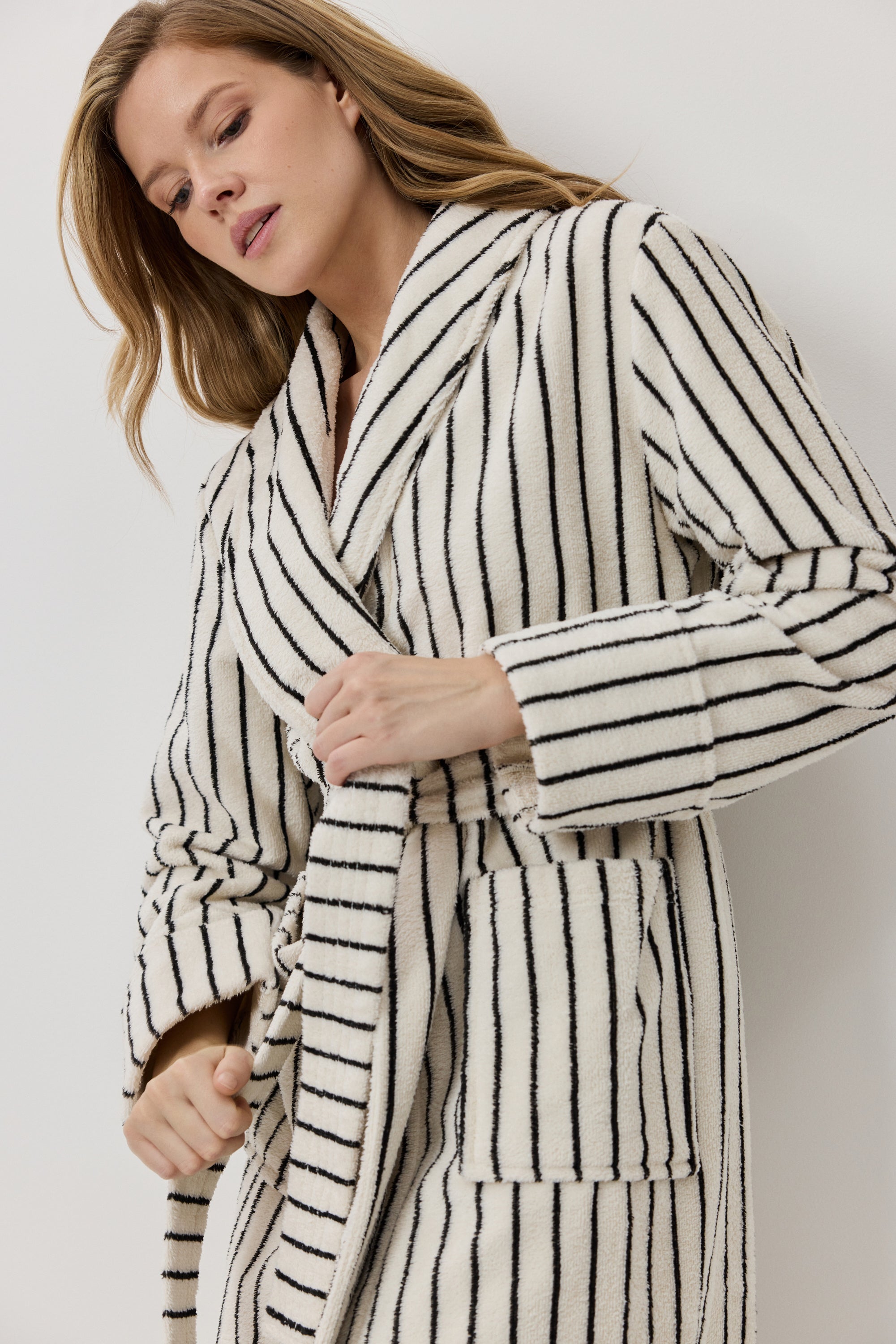 A woman wearing a plush white bathrobe with black vertical stripes, shown in a close-up shot against a light gray background.