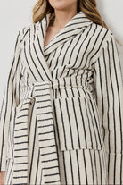 A woman wearing a cream-colored bathrobe with thin black vertical stripes, photographed against a neutral light gray background.